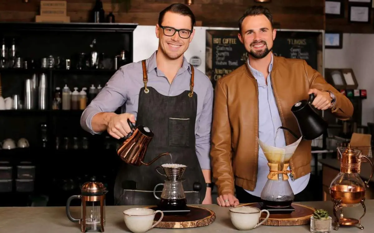 喫茶店でフィルターコーヒーを注ぎながらカメラを覗き込む2人の男性。